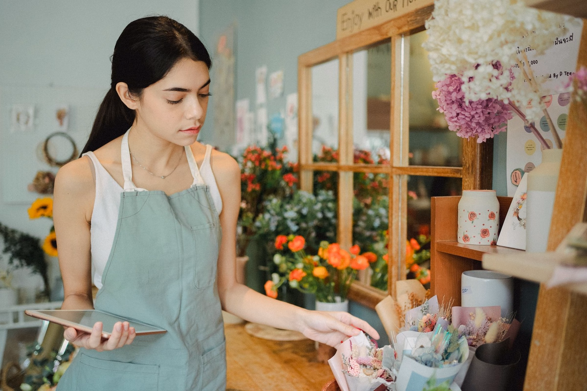 Florist in a green apron holding a tablet while checking bouquets on a shop shelf