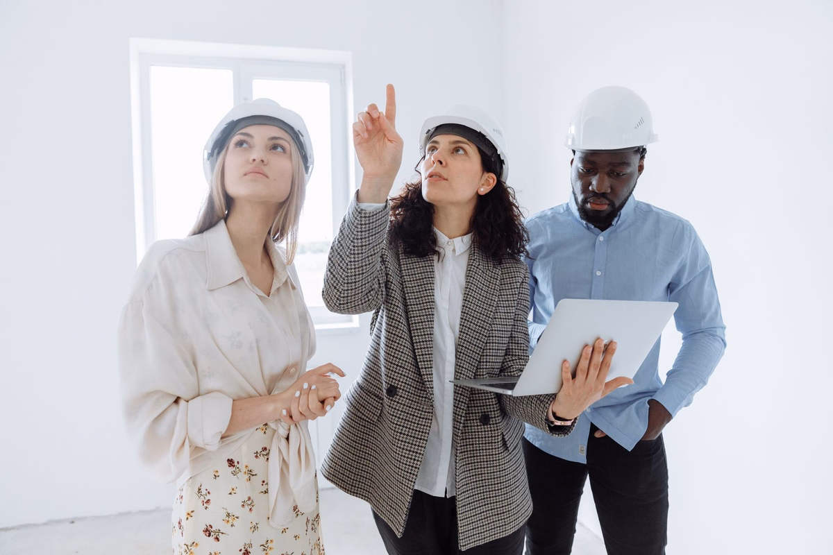 Three people in white hard hats walking through a bright, empty room under renovation, one holding a laptop while another points at the ceiling
