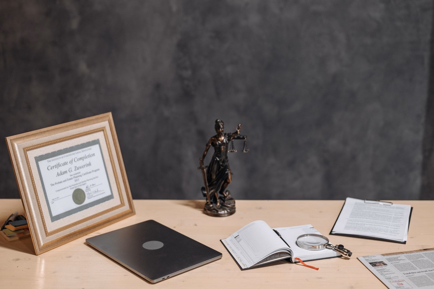 Attorney's desk with a framed certificate, a Lady Justice figurine, a closed laptop, an open notebook with a magnifying glass, and printed documents